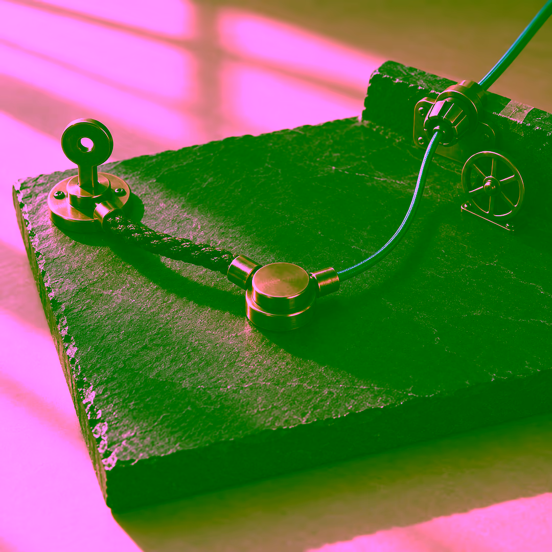 A slate slab in daylight carrying two separate connections to one central round brass socket: on the left a keyed brass token mounted on the slab with a short braided brown cord running to the socket, and from the upper right a teal sleeved cord entering through a collar fitting on the slab's raised edge, with a small spoked wheel mounted beside that collar.