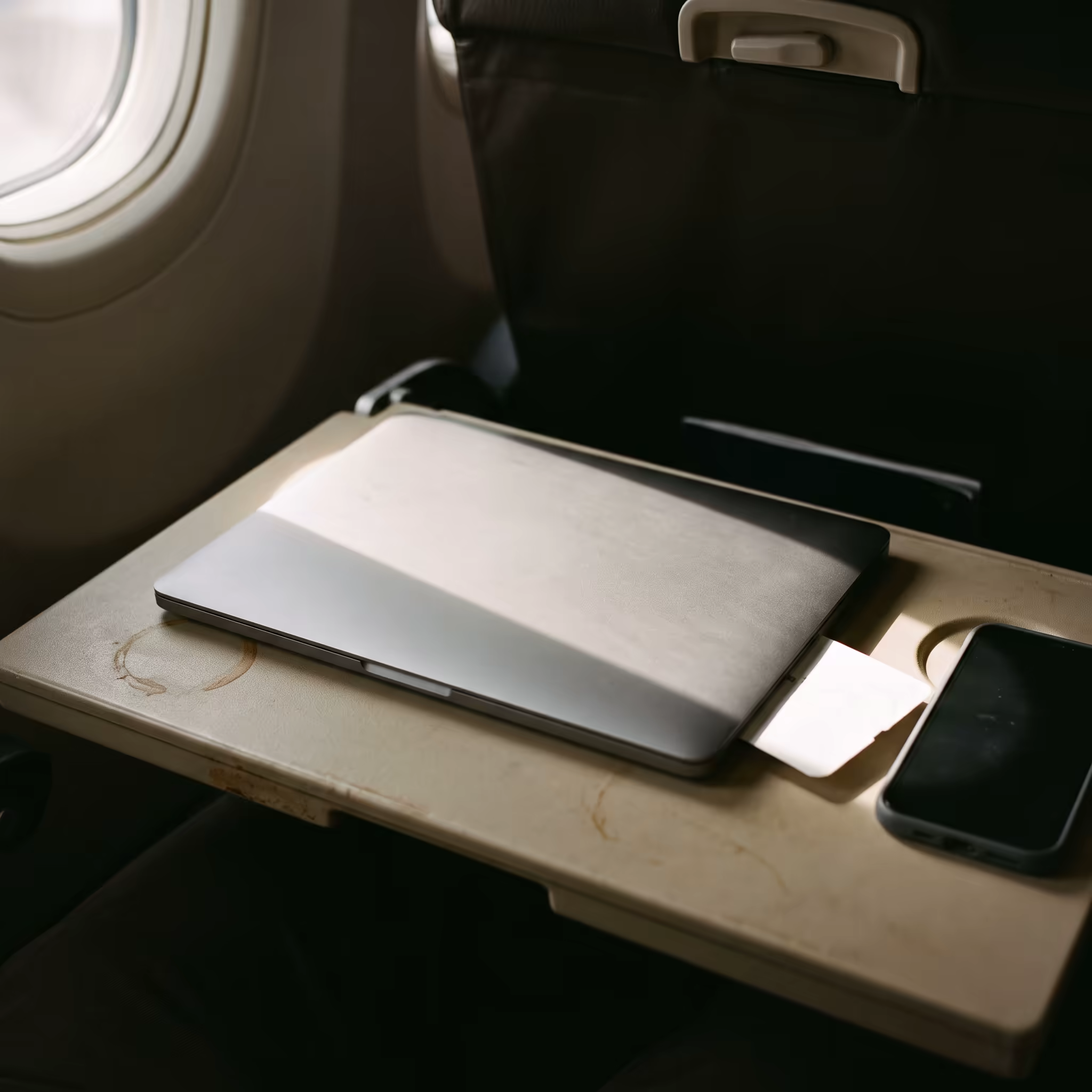 A closed grey laptop on an airline tray table with a hard rectangle of window light falling across the lid, a face-down phone and a dried coffee ring beside it